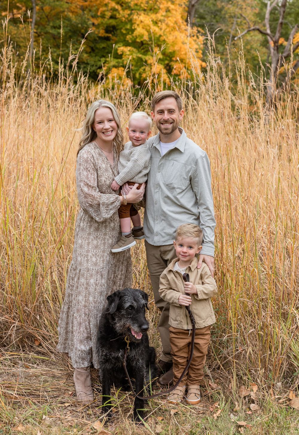 A family poses together in tall golden grass during autumn with their black dog while wearing earth-toned clothing.
