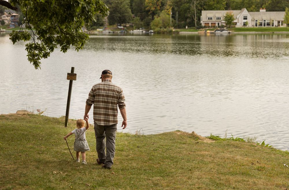 A person and small dog walk along a grassy shoreline beside a tranquil lake on an autumn day.