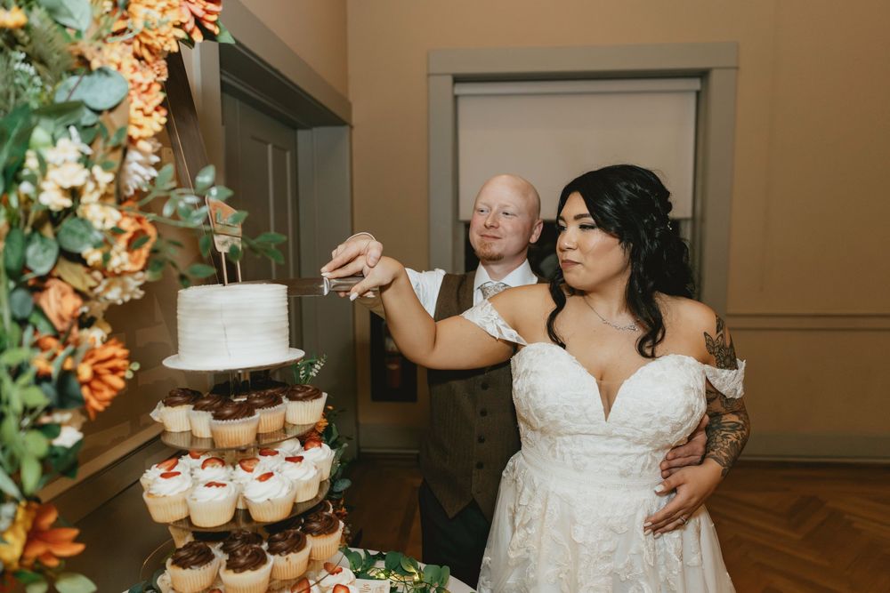 A wedding couple feeds each other cake during their reception while standing next to a white tiered wedding cake.