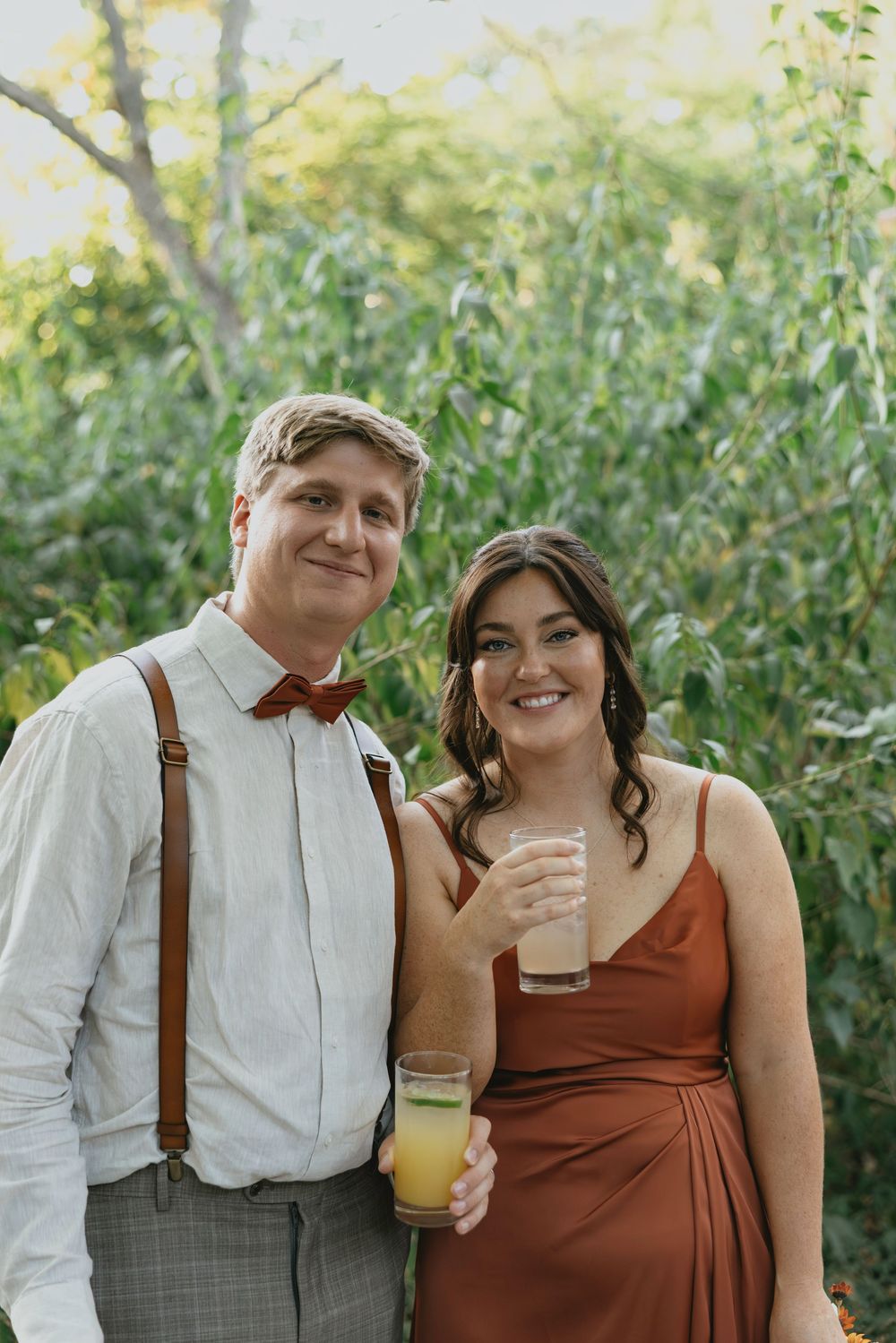 Couple in rustic wedding attire enjoying drinks outdoors with suspenders and brown dress details.