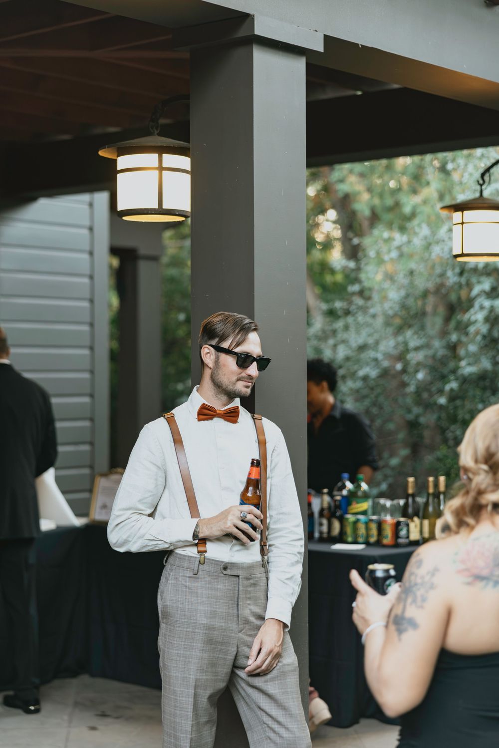 Stylishly dressed guest in suspenders and bow tie enjoys a drink during an outdoor reception.