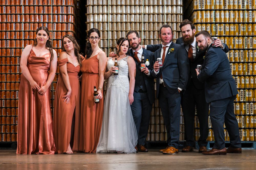 Wedding party members stand in a row against a wooden wall with warm lighting at an indoor venue.