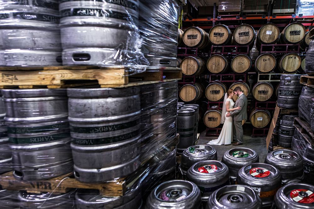 Rows of metal beer kegs stacked in a brewery warehouse with wooden barrels visible in the background.