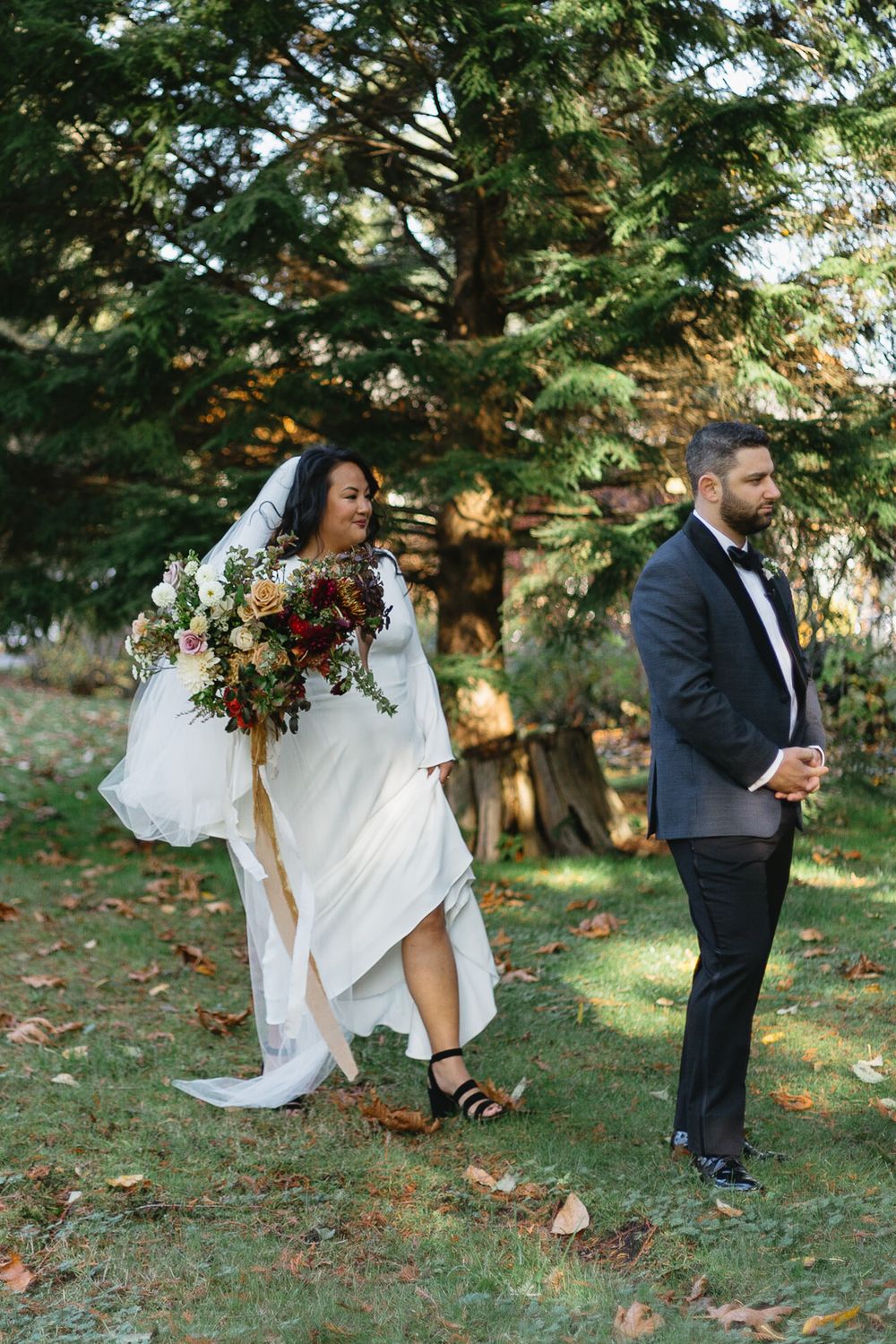 A wedding couple playfully searches for something in fallen autumn leaves beneath evergreen trees in a park setting.