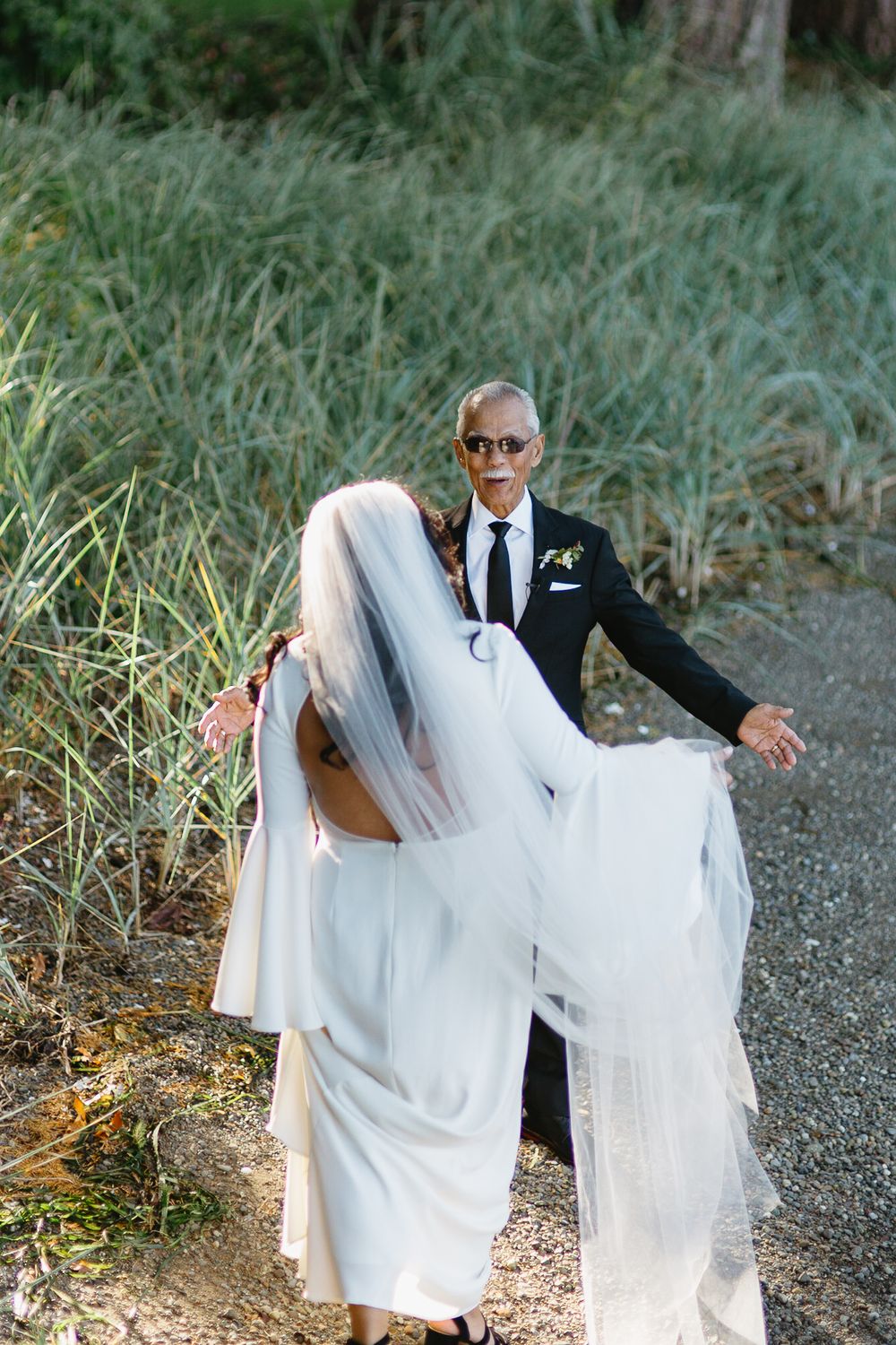 A couple in wedding attire poses together on a gravel path with tall grass and greenery in the background.