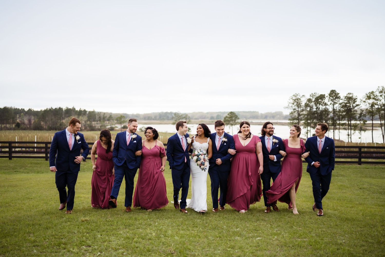 Wedding party members in burgundy dresses and dark suits pose together on a grassy field near a fence.