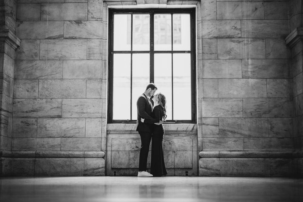 Black and white portrait of couple by large window in stone building.