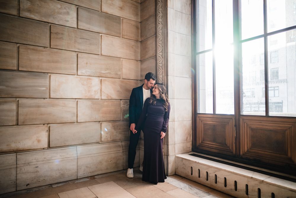 Couple stands close together by large window in stone hallway.