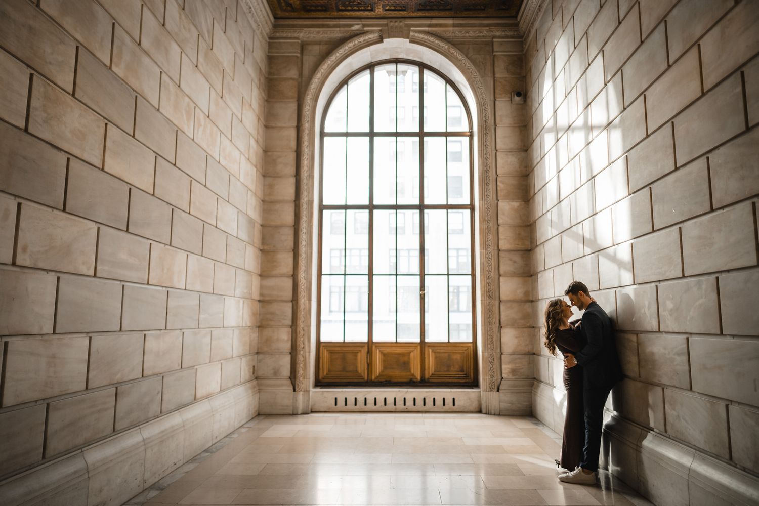 Couple embraces in sunlit stone corridor with tall arched window.