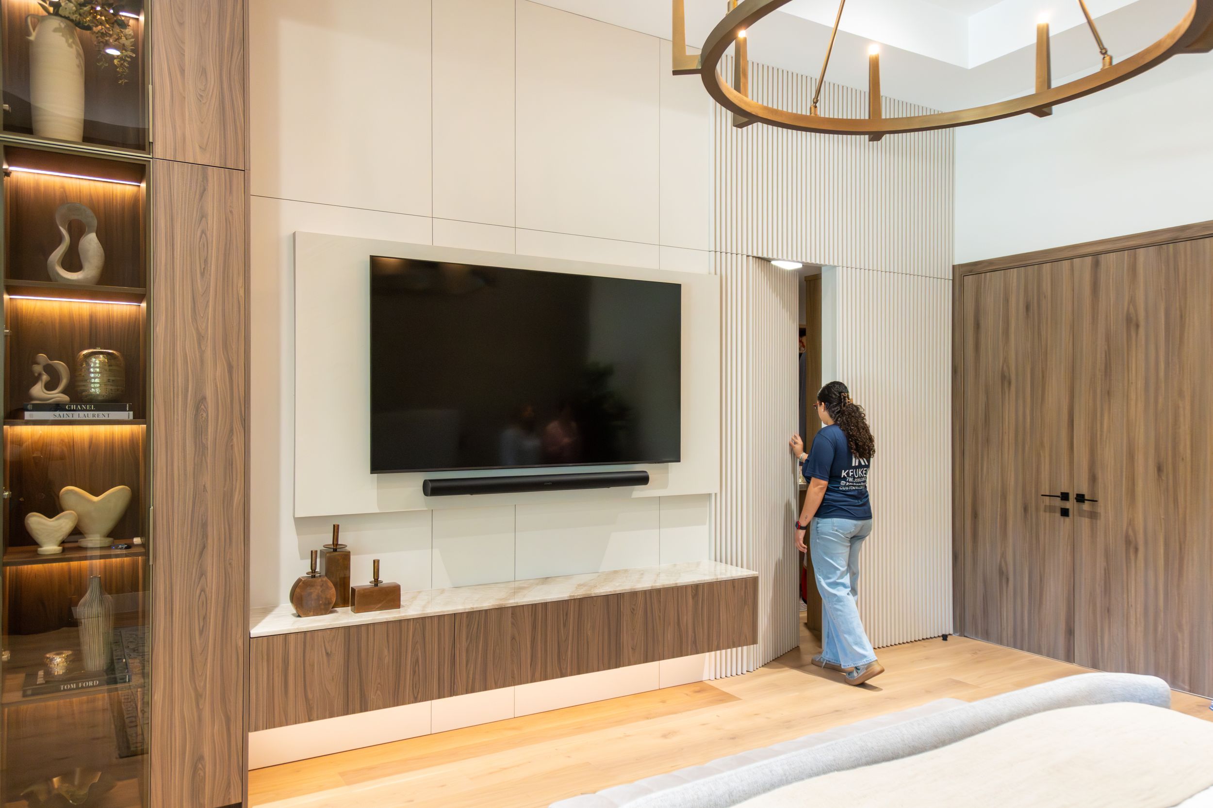 Woman standing in a modern bedroom with a large wall-mounted TV, wooden accents, and a circular chandelier overhead.