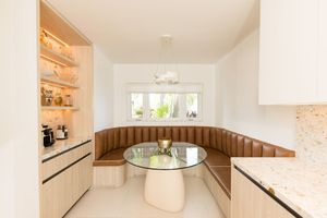 Modern breakfast nook with curved tan leather banquette, round glass table, built-in shelving, and white cabinetry.