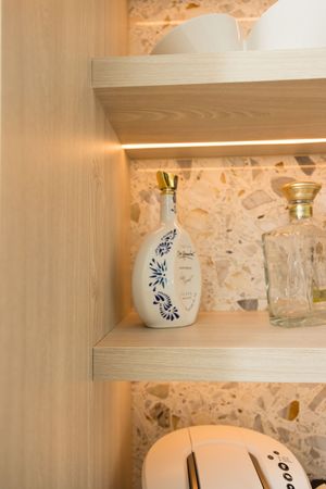 Modern built-in wooden shelves with LED lighting displaying decorative bottle, glassware, and dishes against stone tile wall.