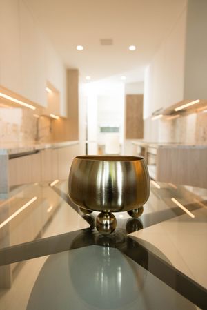 Gold decorative bowl on glass table in modern luxury kitchen with white cabinets and recessed lighting.