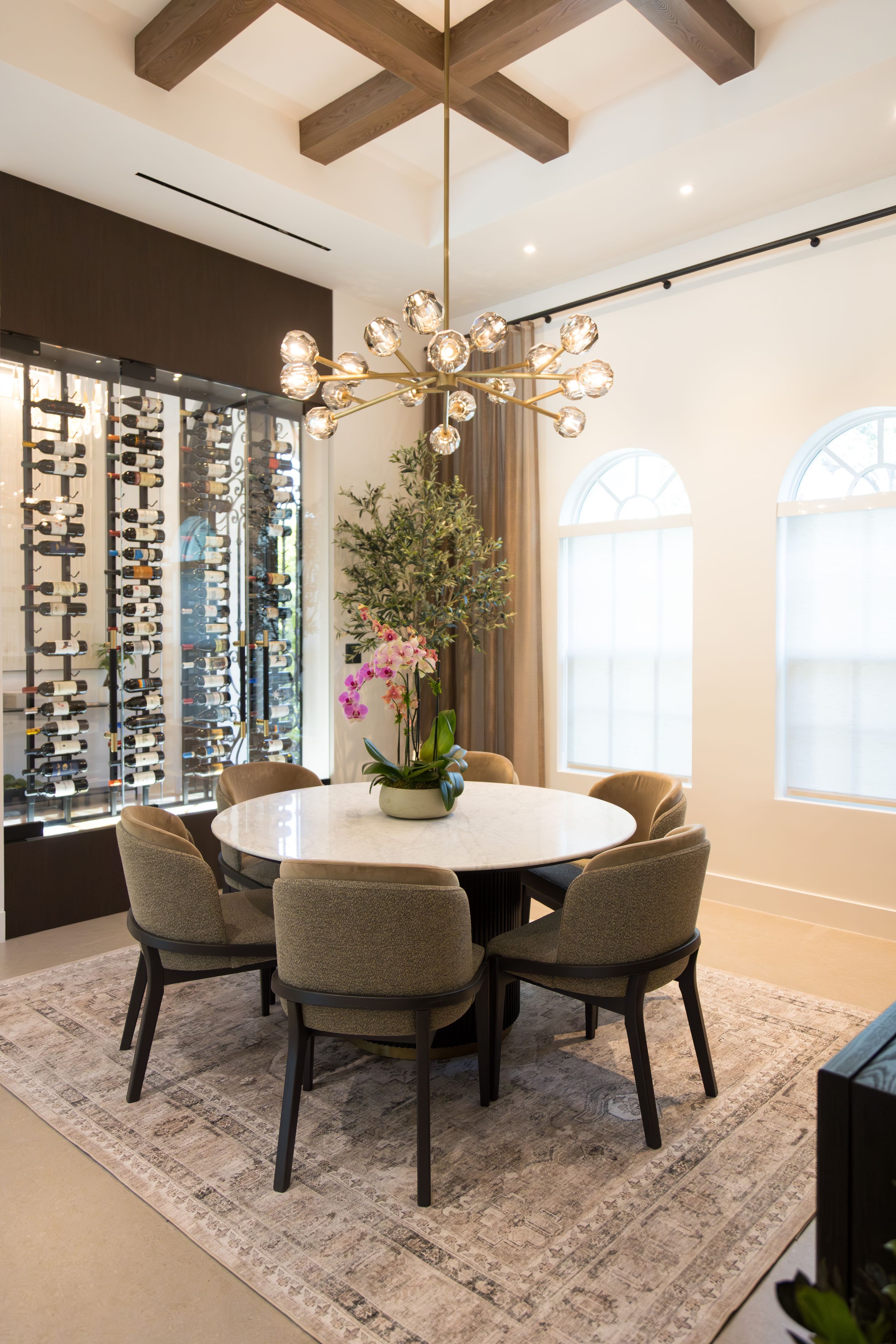Elegant dining room with round white table, gray chairs, modern chandelier, wine wall, and arched windows on beige rug.