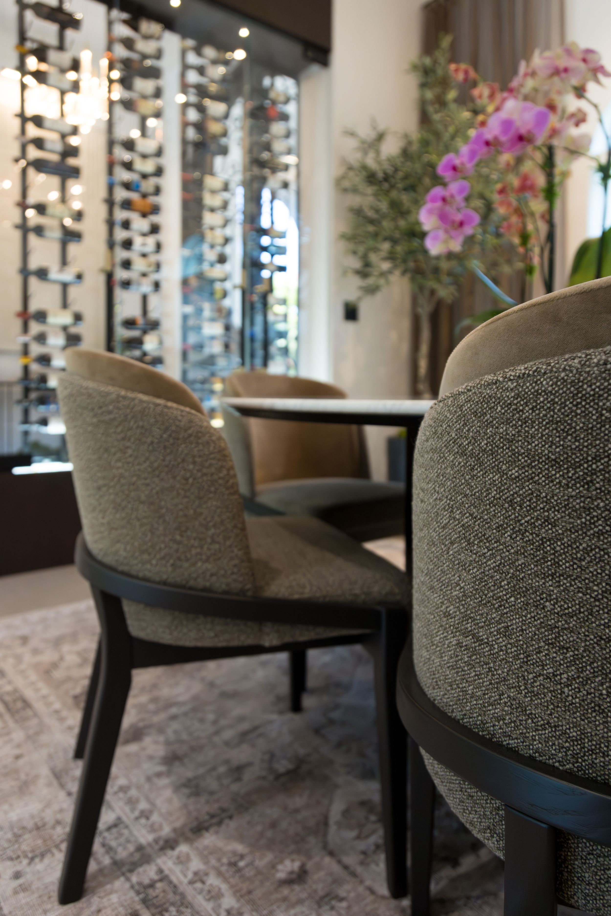 Elegant dining area with upholstered chairs, round table, wine rack wall display, and pink orchids in a modern restaurant.