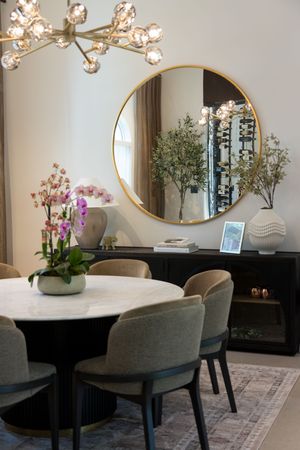 Elegant dining room with marble table, gray chairs, gold round mirror, black sideboard, orchid centerpiece, and modern chandelier.