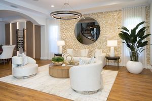Elegant living room with white curved sofas, gold accents, round mirror, patterned wall, and lush plant on hardwood floors.
