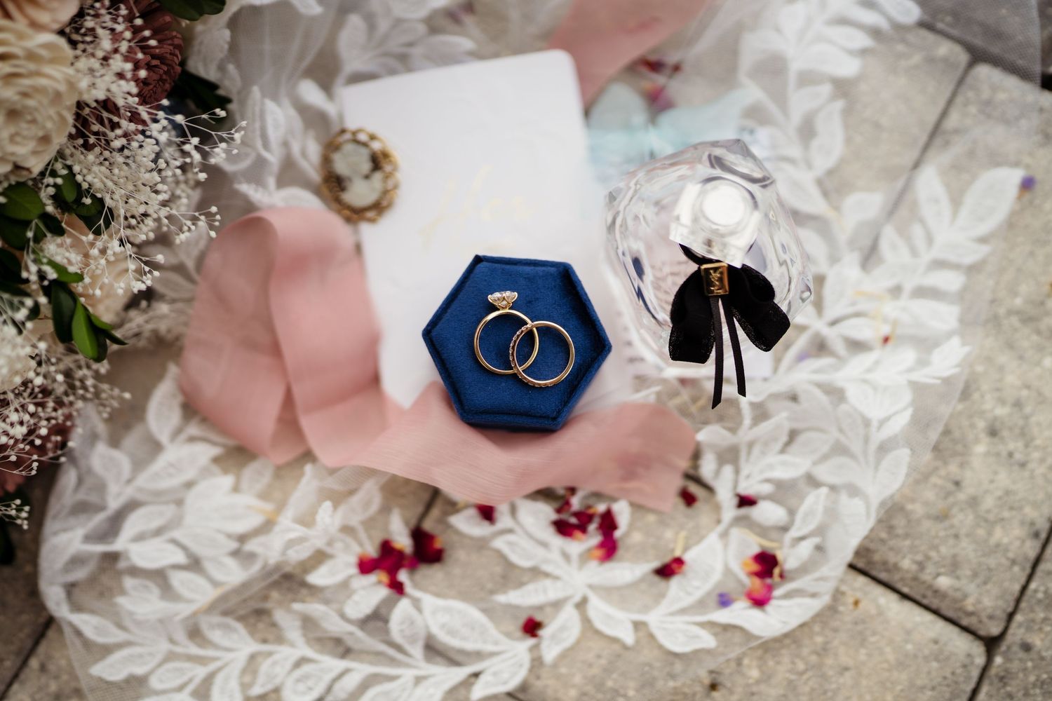 Wedding rings and decorative elements arranged on white lace with scattered red rose petals.