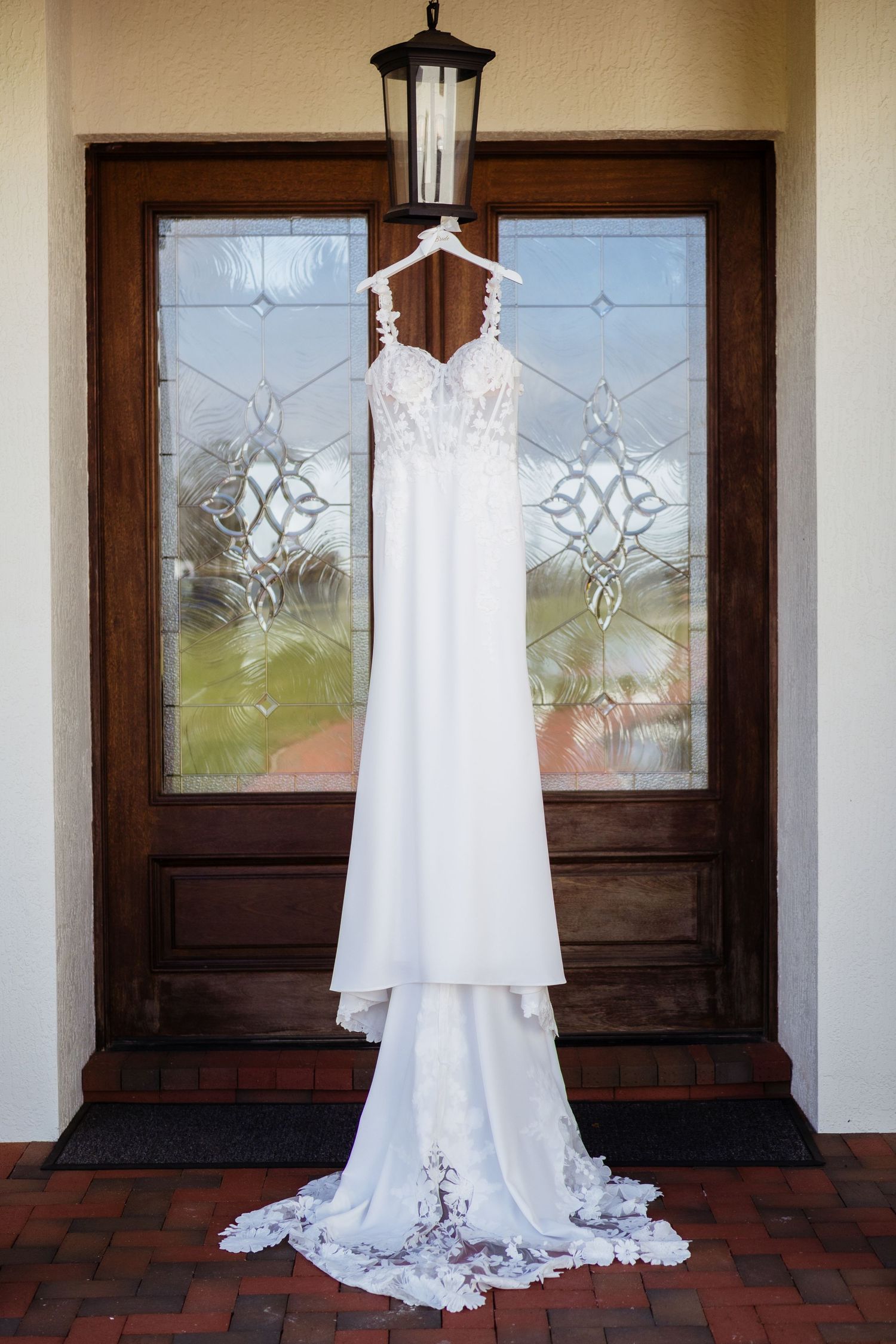 A beautiful white wedding dress hangs elegantly against dark wooden doors with decorative glass panels.