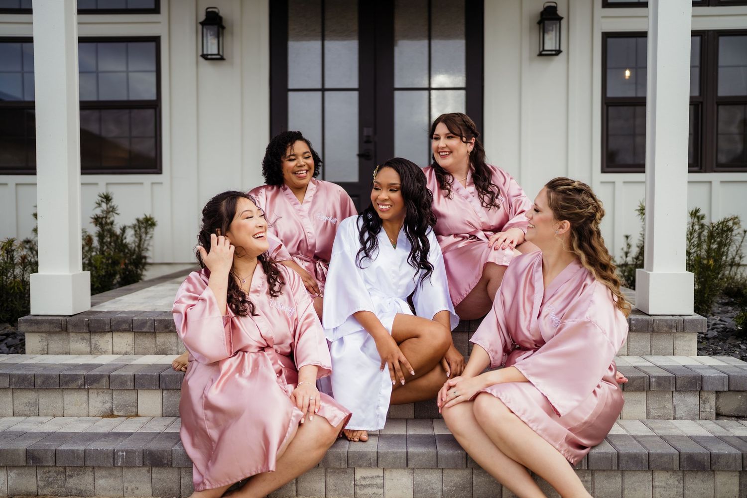 Group wearing matching pink satin robes and white tops sit laughing together on outdoor steps.