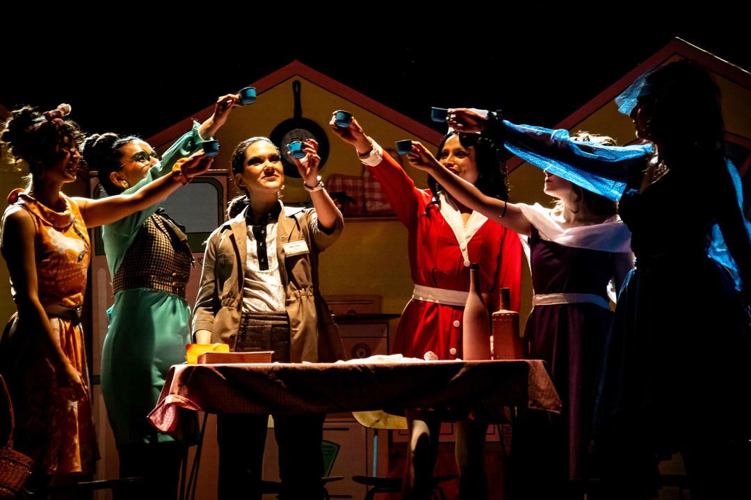 Dramatic theater scene with performers in period costumes gesturing around a table during an intense stage performance.