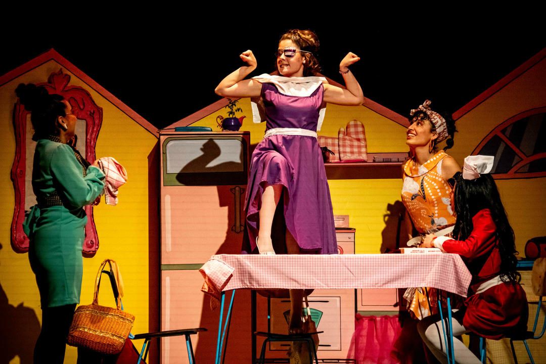 Theatrical performers act out a scene on a colorful stage set designed to look like the interior of a house.