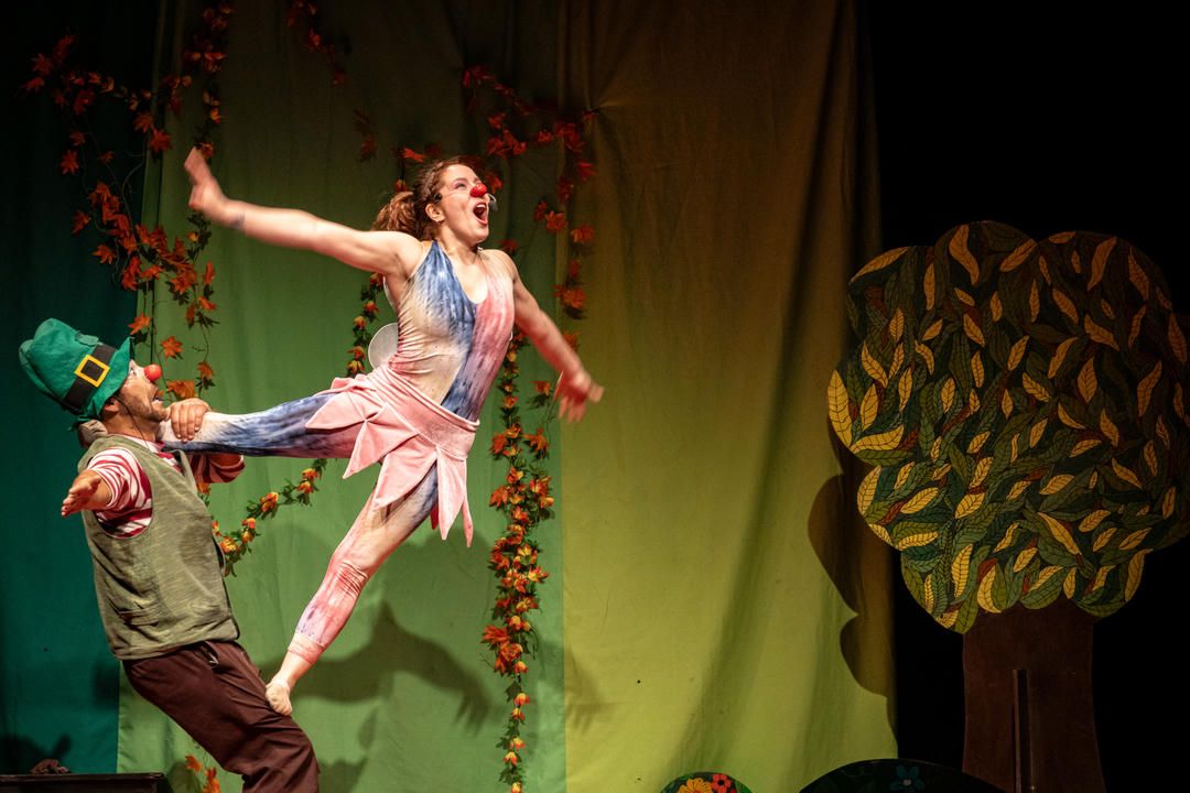 Performers in colorful costumes dance and interact on a green-lit stage with hanging vines during a theatrical production.
