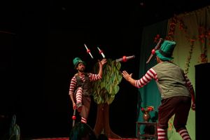 Performers in colorful costumes dance and interact on a dimly lit stage during a theatrical production.