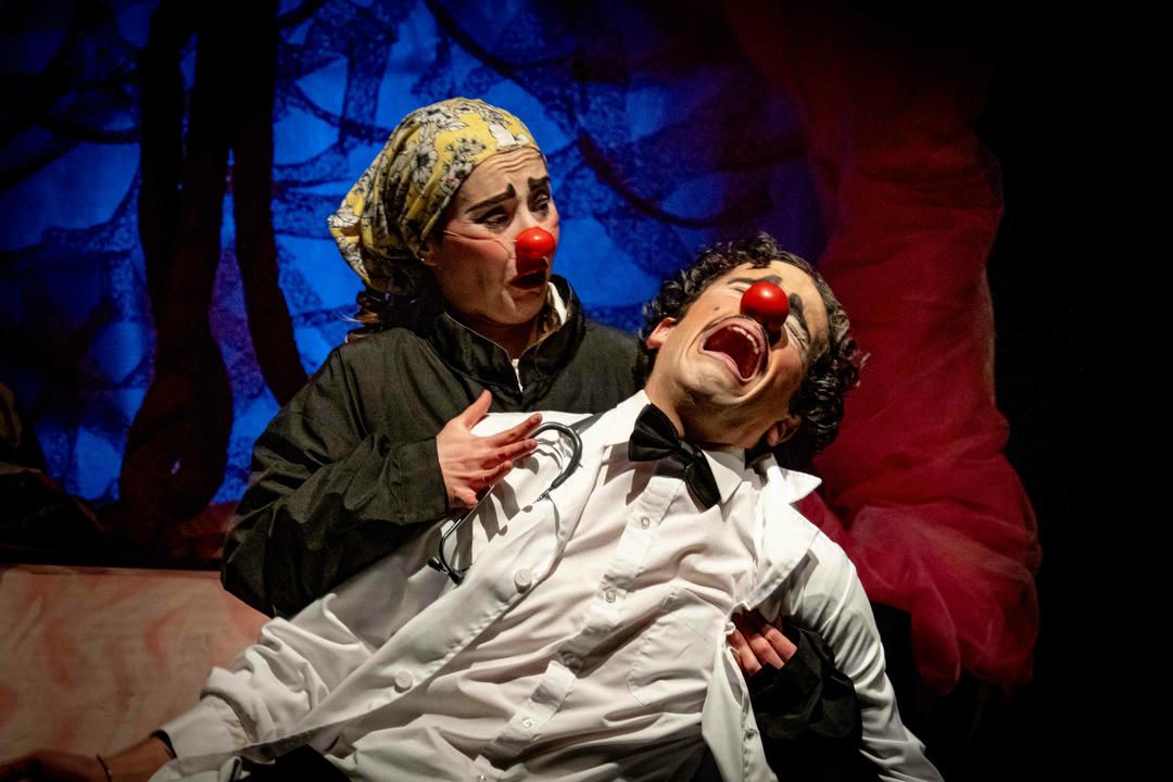 Two performers in white costumes engage in a dramatic scene with red and blue theatrical lighting.