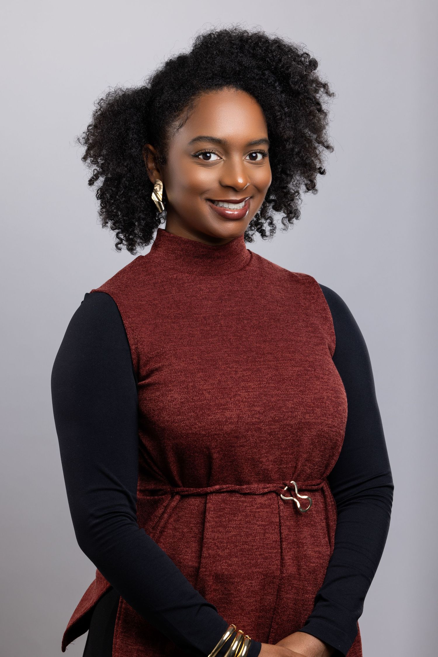 Smiling model in a burgundy turtleneck dress with black sleeves poses against a gray backdrop.