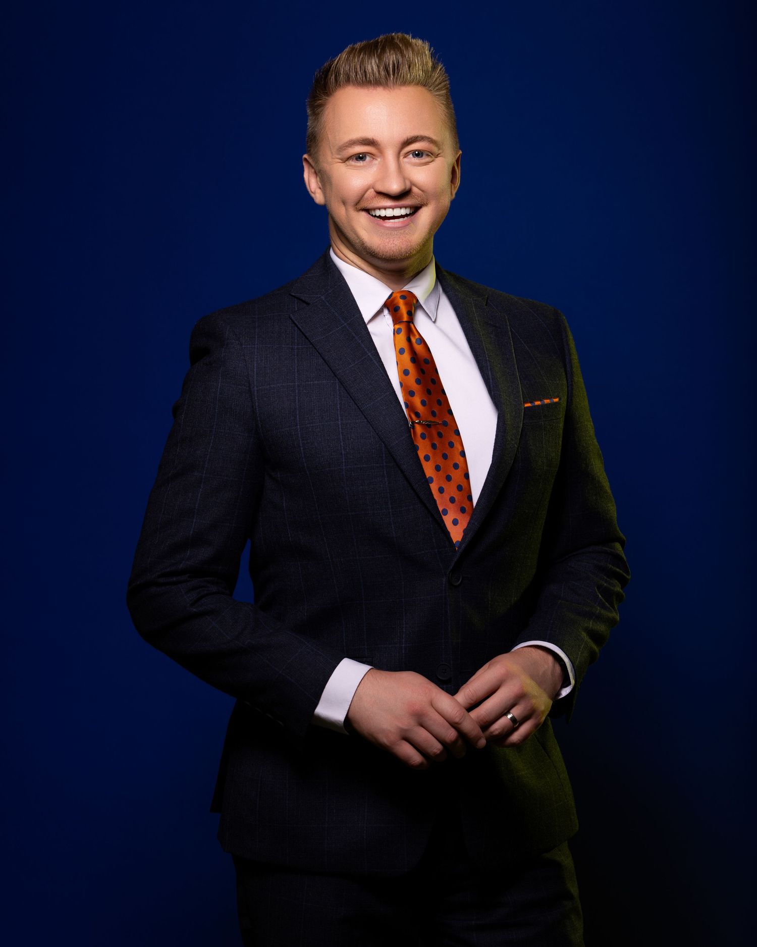 Professional business portrait of someone in a dark suit and red patterned tie against a navy blue background.