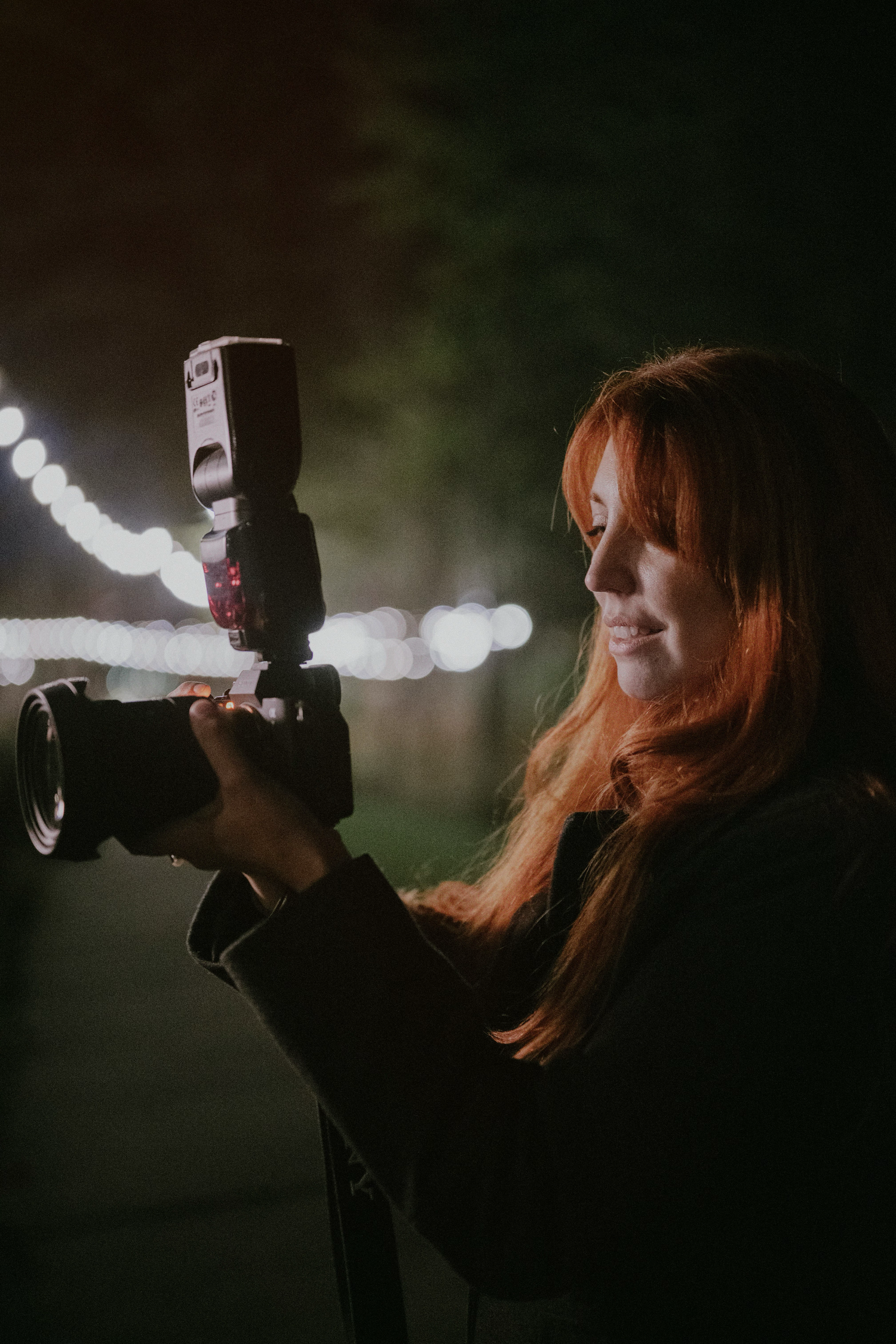 Photographer with vibrant red hair holds camera at night with bokeh lights creating dreamy atmosphere in background.