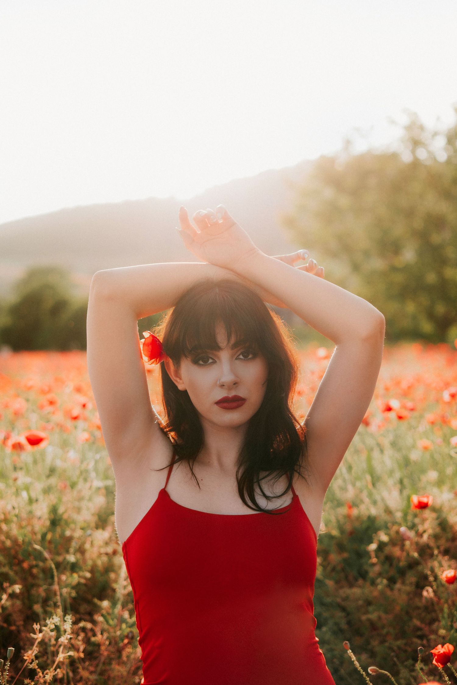 Brunetka v červených šatách na tenkých ramienkach s rukami zdvihnutými nad hlavou pózuje v makovom poli počas zlatej hodinky. Fotografia,