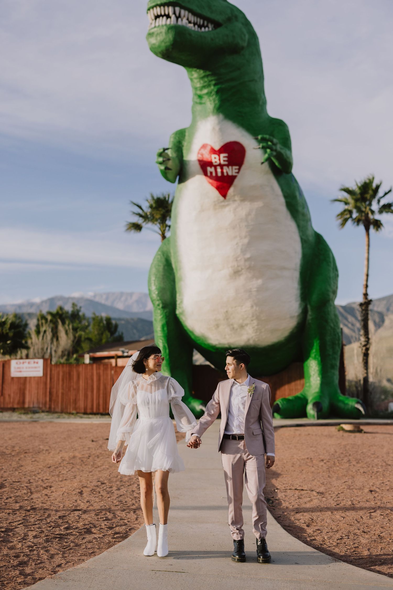 Couple holds hands walking in front of a giant T-Rex dinosaur statue in the desert.