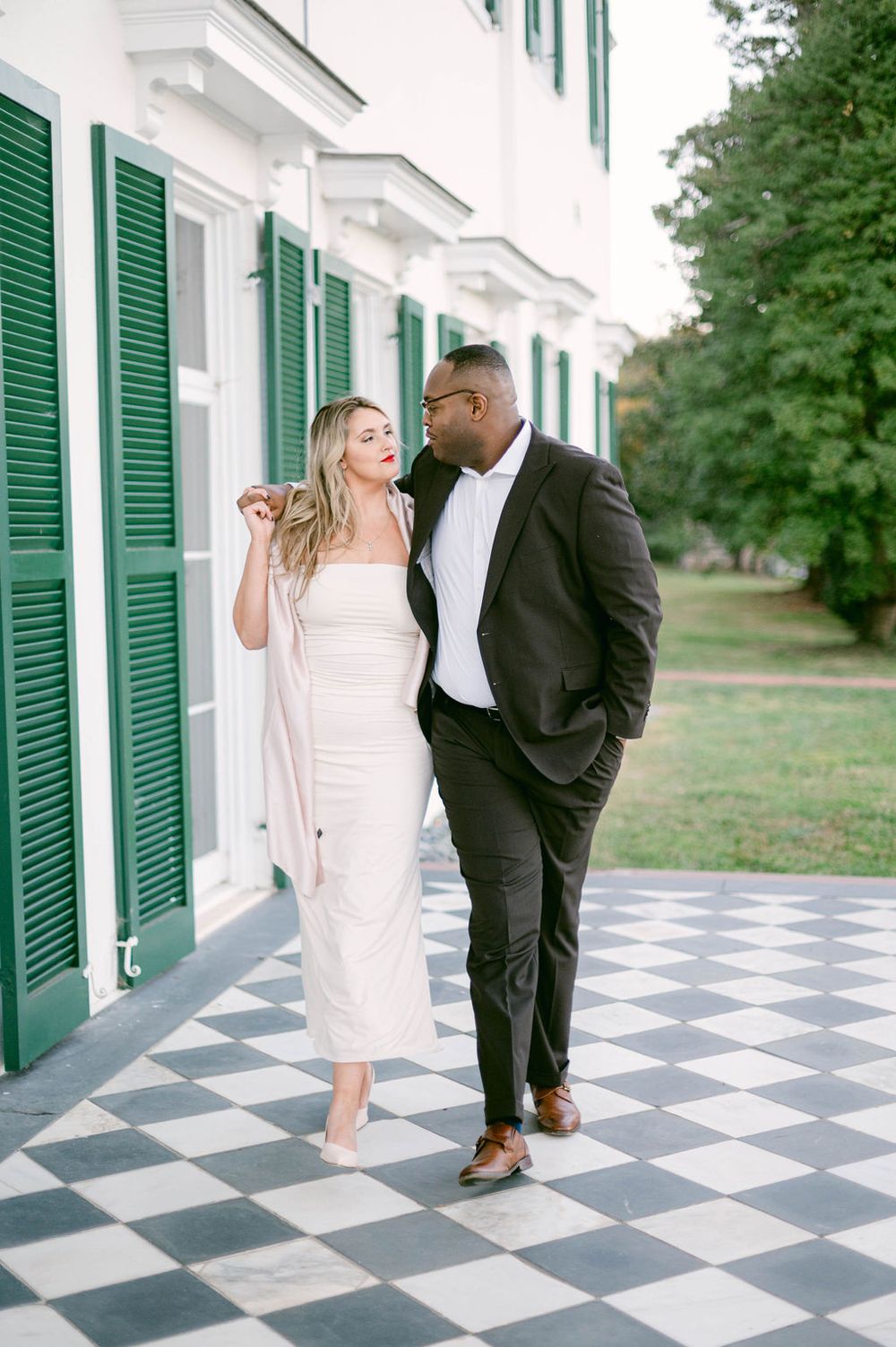 A couple dances playfully on a black and white checkered porch with green shuttered windows behind them.