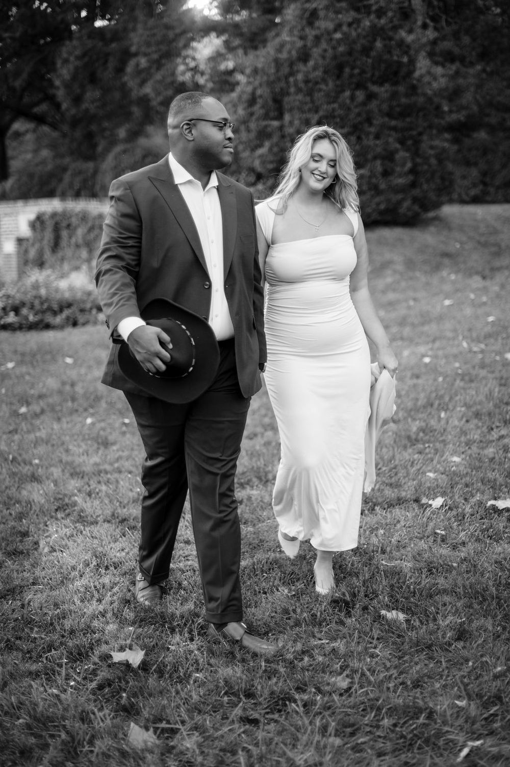 A couple walking together in a garden, with the man wearing a dark suit and holding a hat in black and white photography.