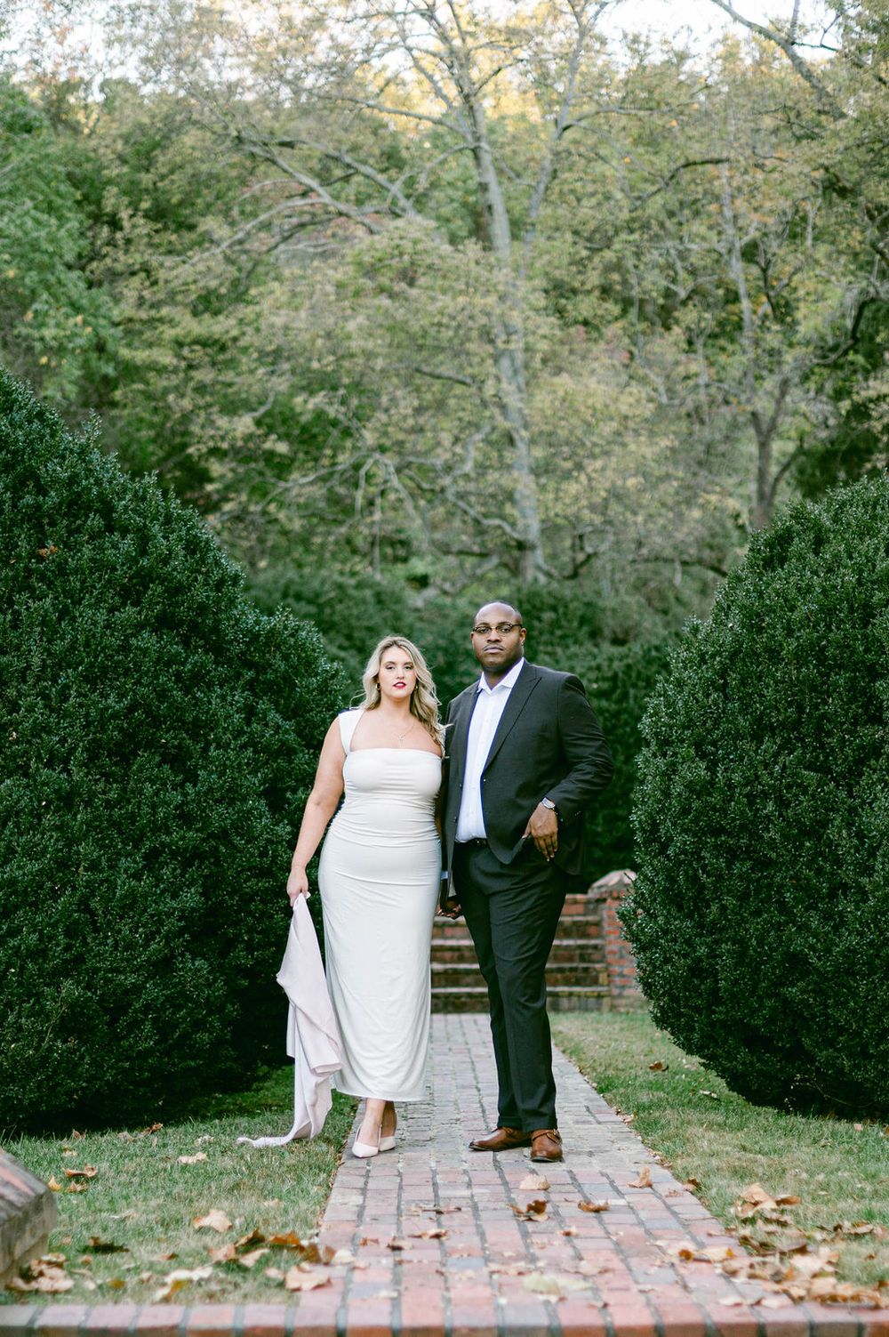 A couple walks together on a brick pathway lined with green hedges and fallen autumn leaves.