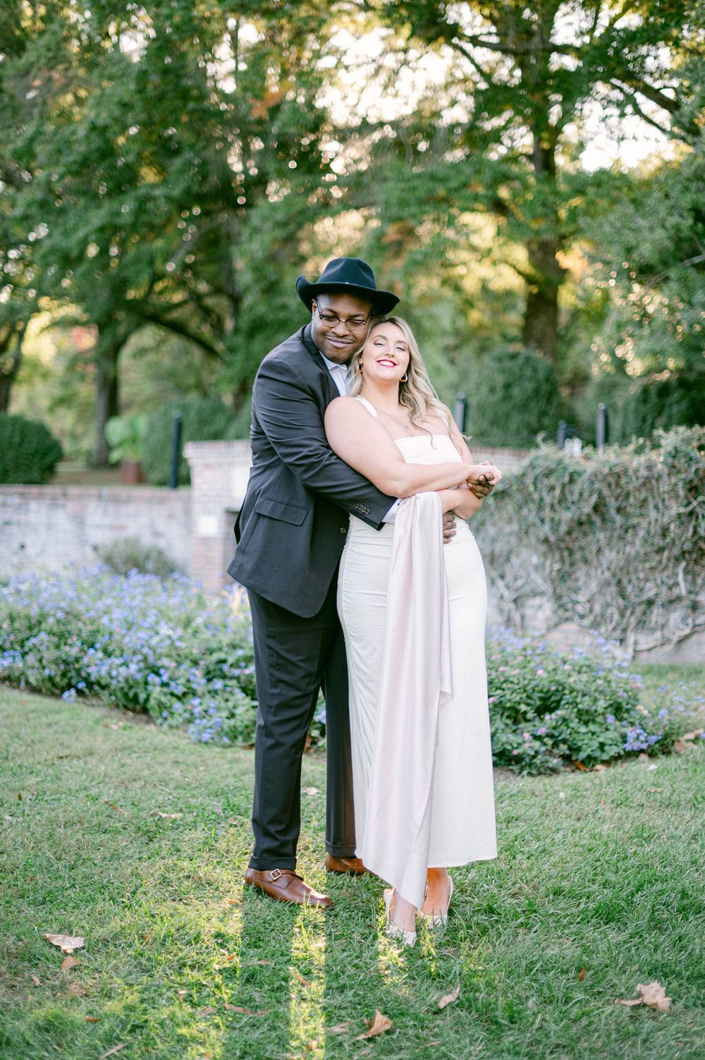 A couple in formal attire shares a romantic dance moment together on a grassy hillside surrounded by greenery at sunset.
