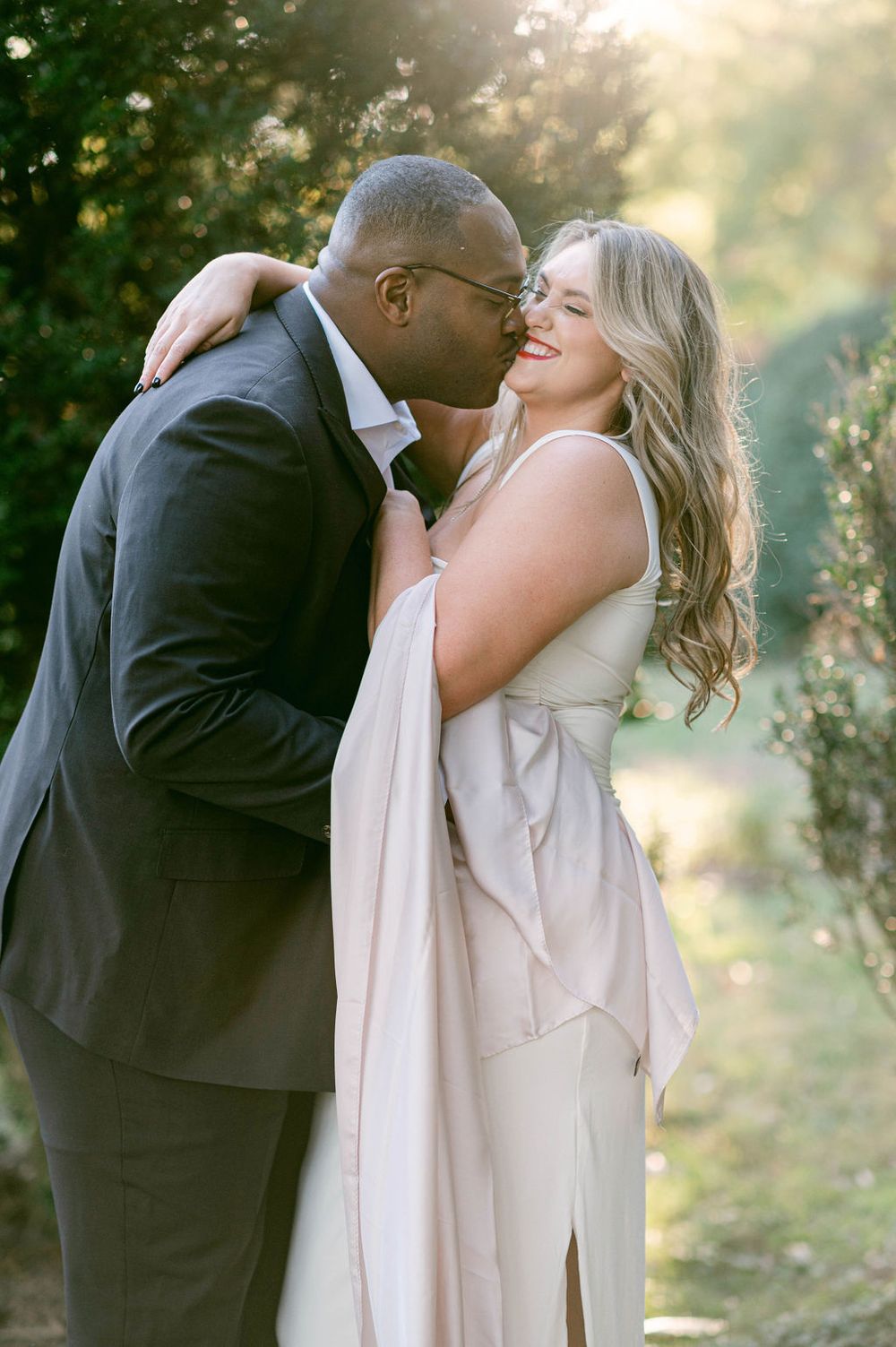 A romantic couple embraces and laughs together in a dreamy outdoor setting with golden sunlight filtering through trees.