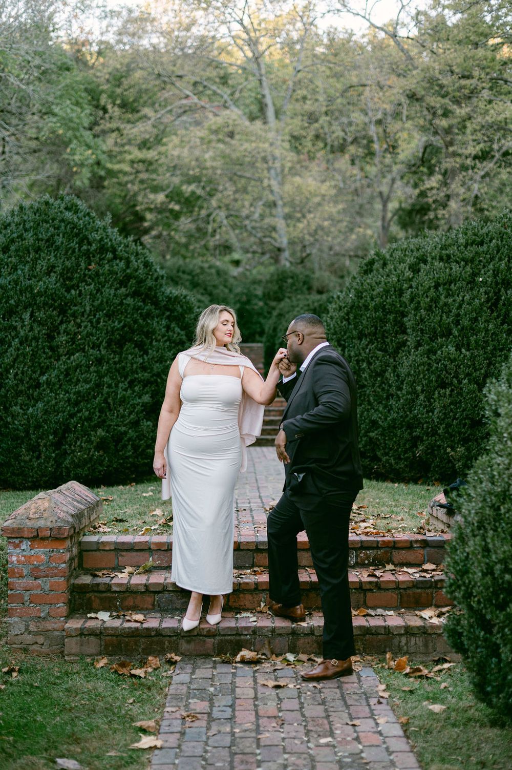 A couple shares a romantic moment on a brick wall pathway surrounded by lush greenery on an autumn day.