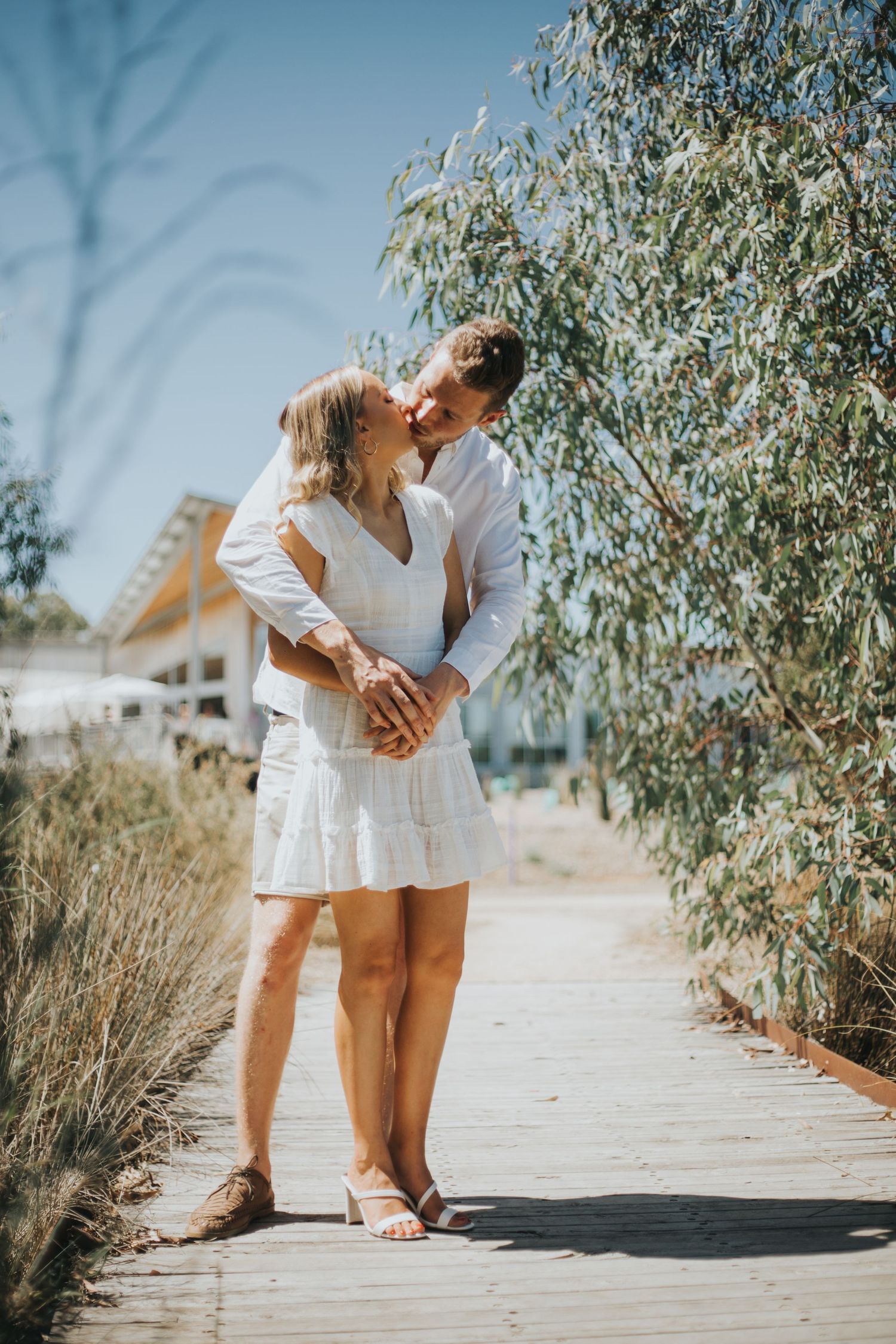 Couple kissing on a wooden boardwalk surrounded by dry grass and eucalyptus trees on a sunny day.