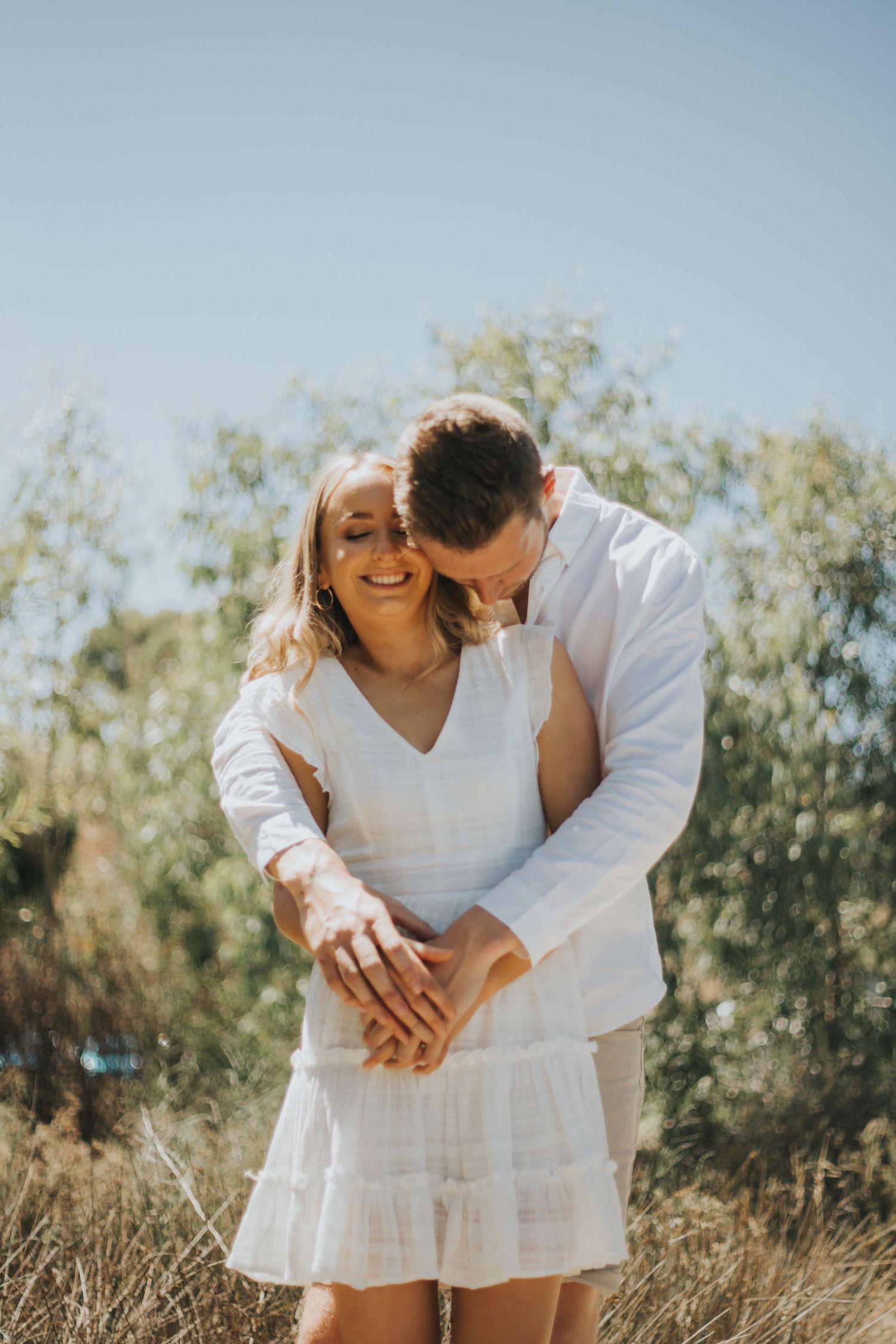 Happy couple embracing outdoors, man kissing womans head while she smiles in a sunny natural setting.