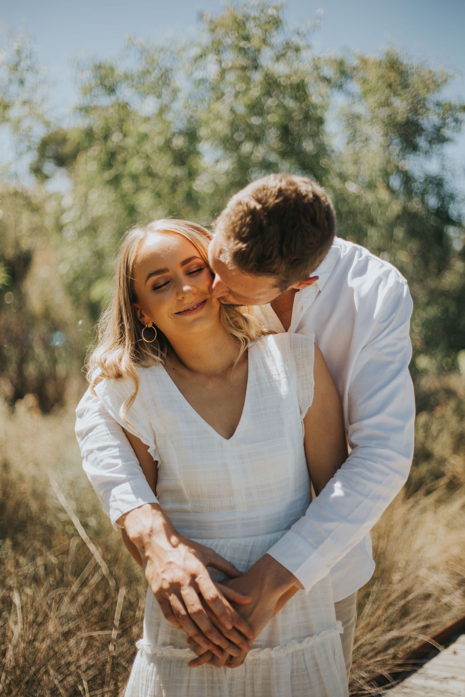 Romantic couple embracing in golden grass, man kissing womans cheek as she smiles with eyes closed.