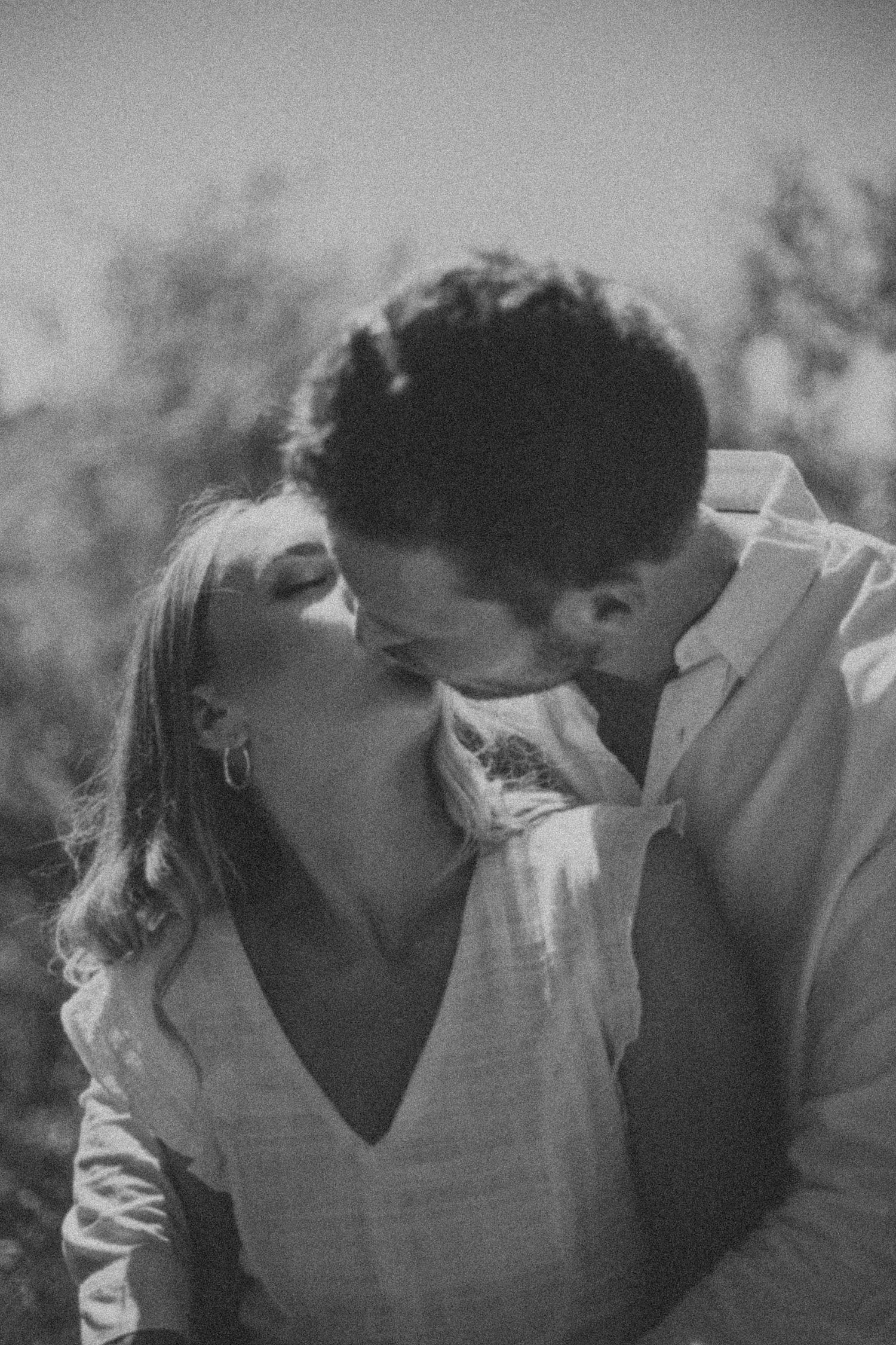 Intimate black and white photo of a couple kissing closely with blurred trees in the background.