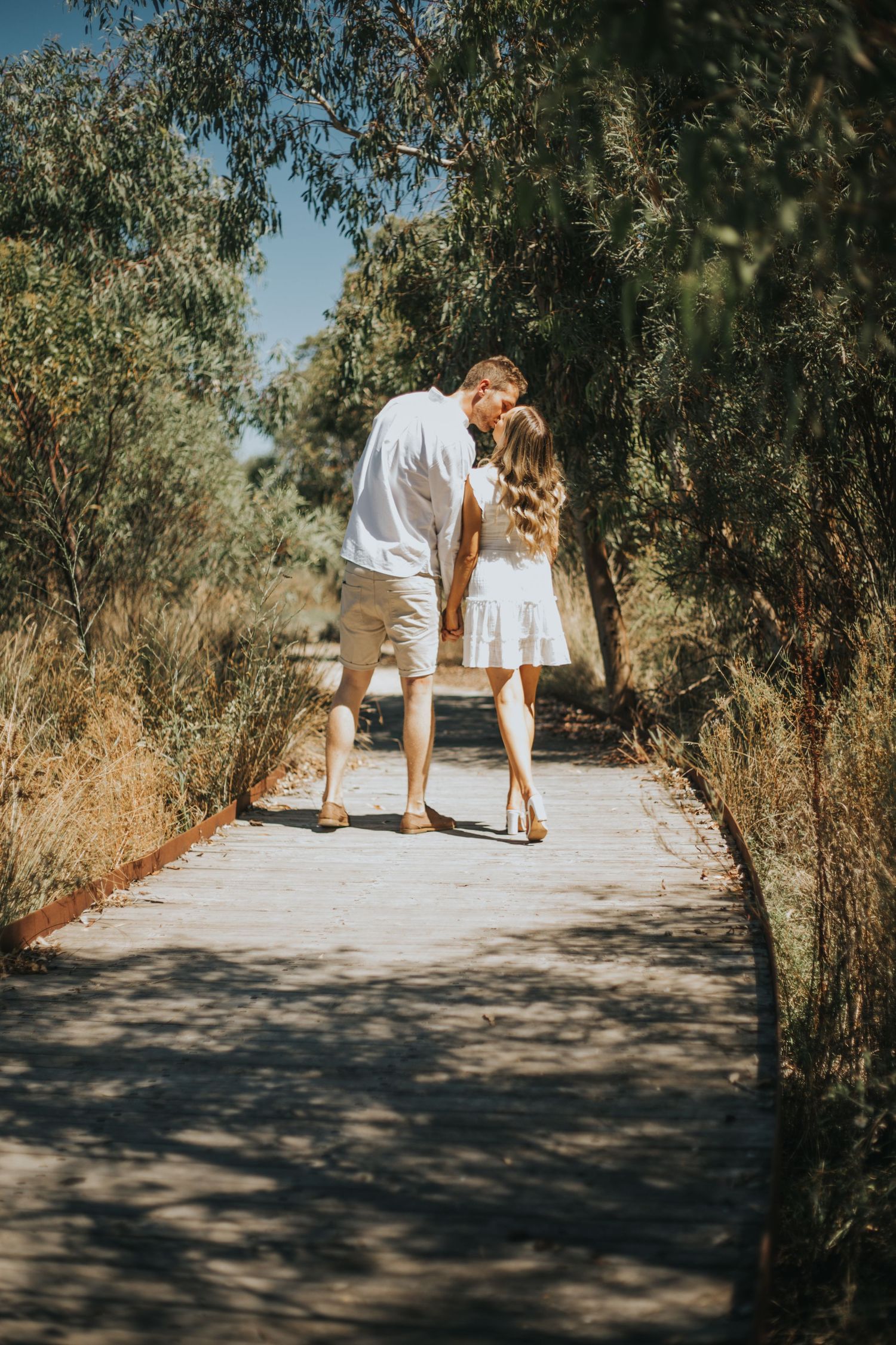 Couple kissing on a sunlit tree-lined pathway, both dressed in white on a bright sunny day.
