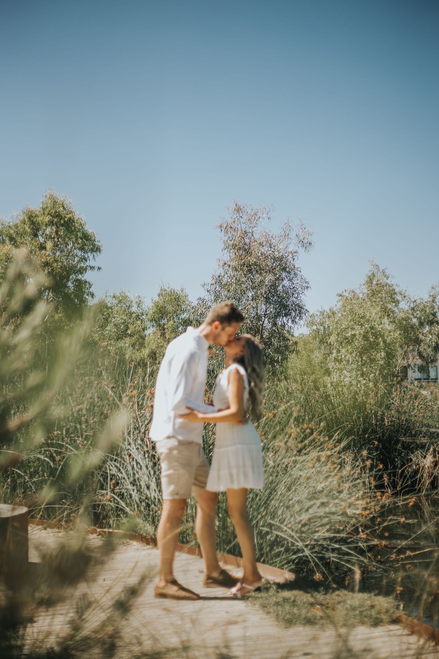 Couple sharing a kiss on a scenic garden path surrounded by lush green shrubs under a clear sky.