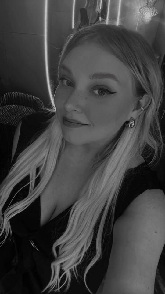 Black and white selfie of a young blonde woman with winged eyeliner and stud earrings smiling softly.