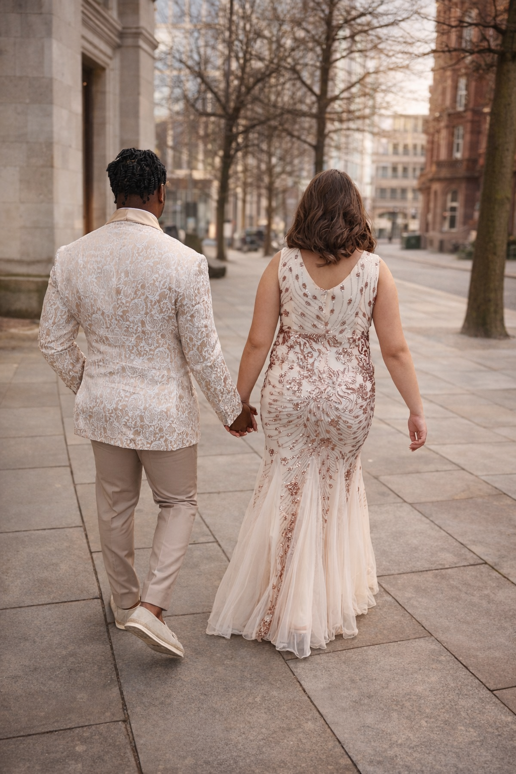 Two people in white lace wedding attire walk together on a stone pathway during sunset at an urban venue.