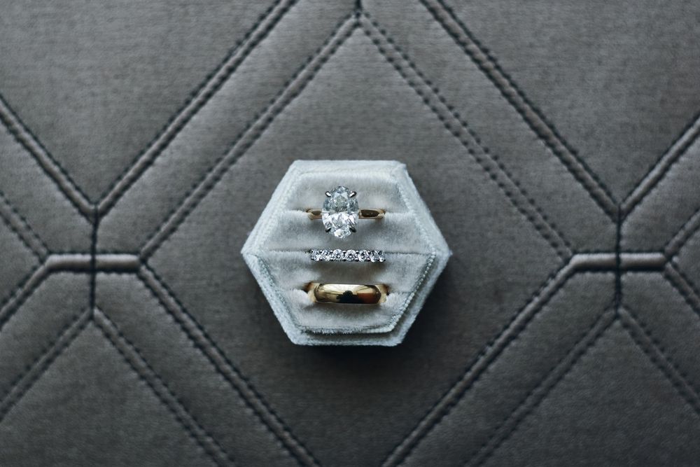 A sparkling diamond engagement ring sits in a hexagonal clear ring box on a gray textured surface.
