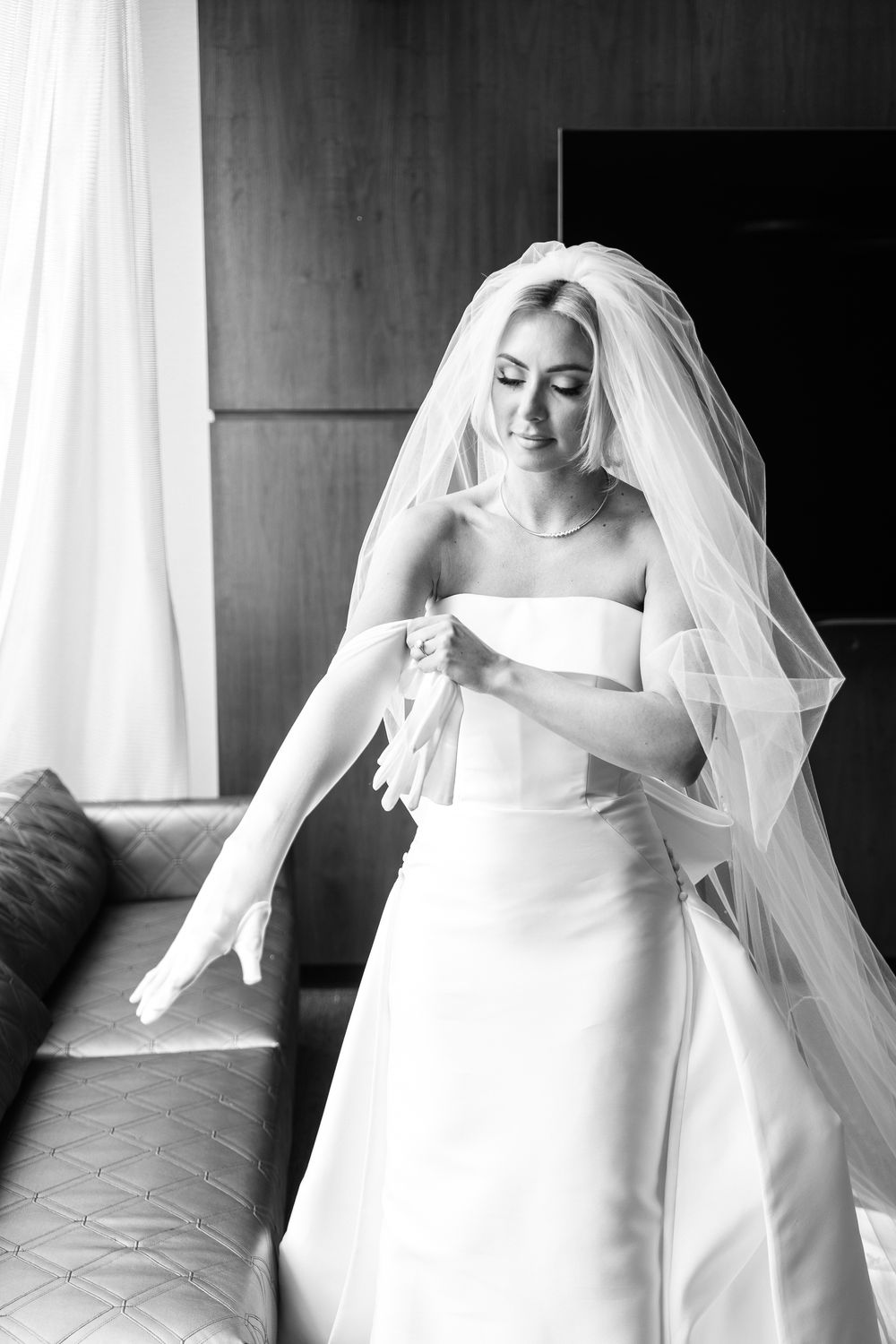 A bride in an elegant white strapless wedding gown and flowing veil stands gracefully in a room with natural light.